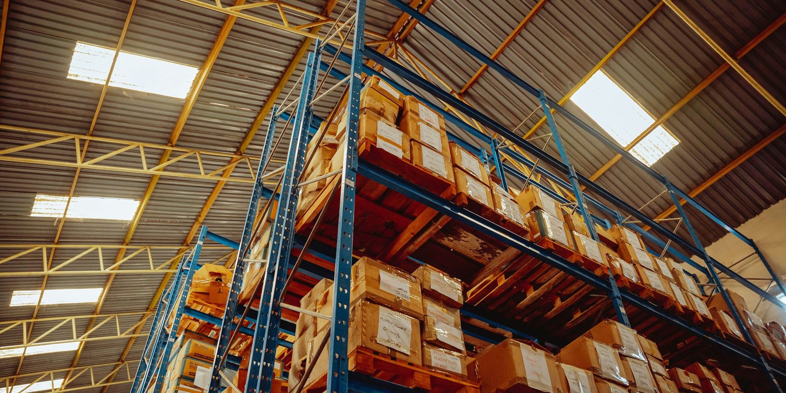 Tall pallet racking with blue uprights and orange beams loaded with boxed goods under a skylit warehouse roof