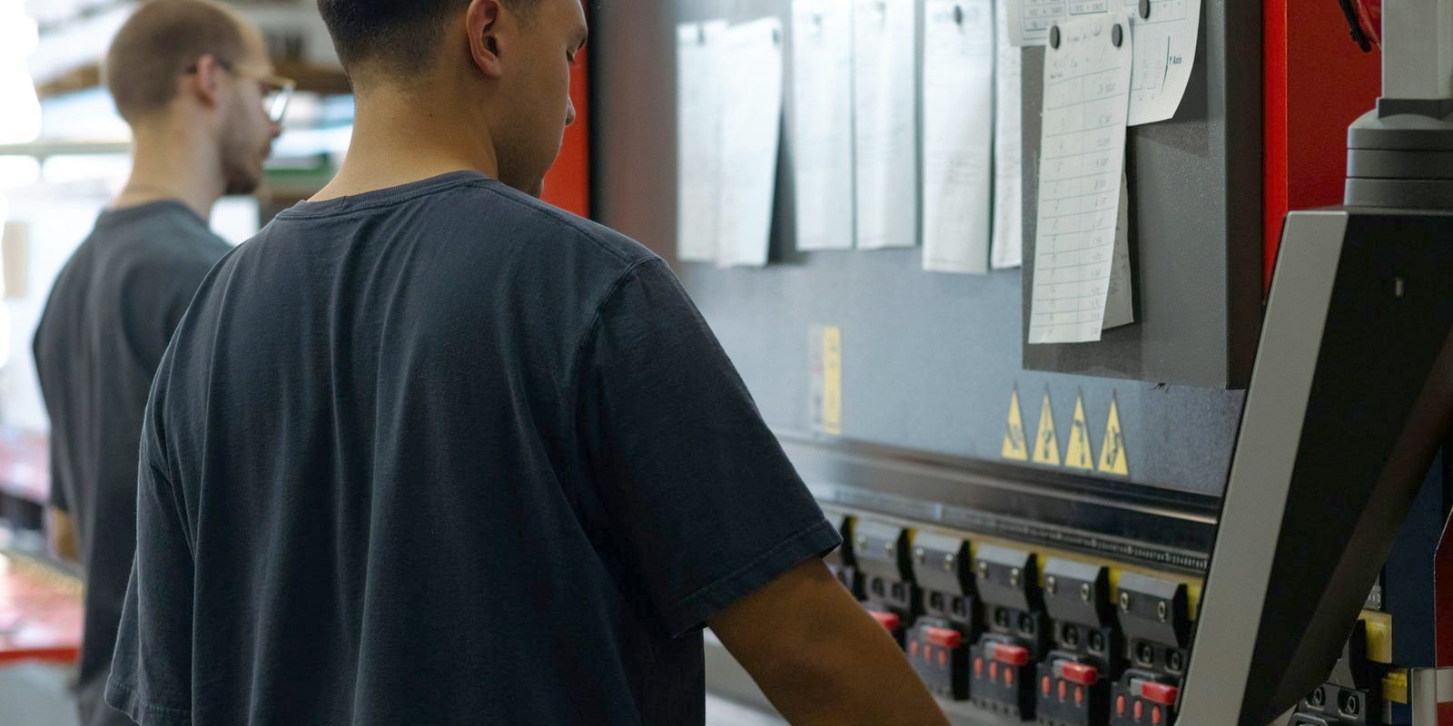 Two operators at a press brake in a sheet metal shop, with job sheets pinned to the machine