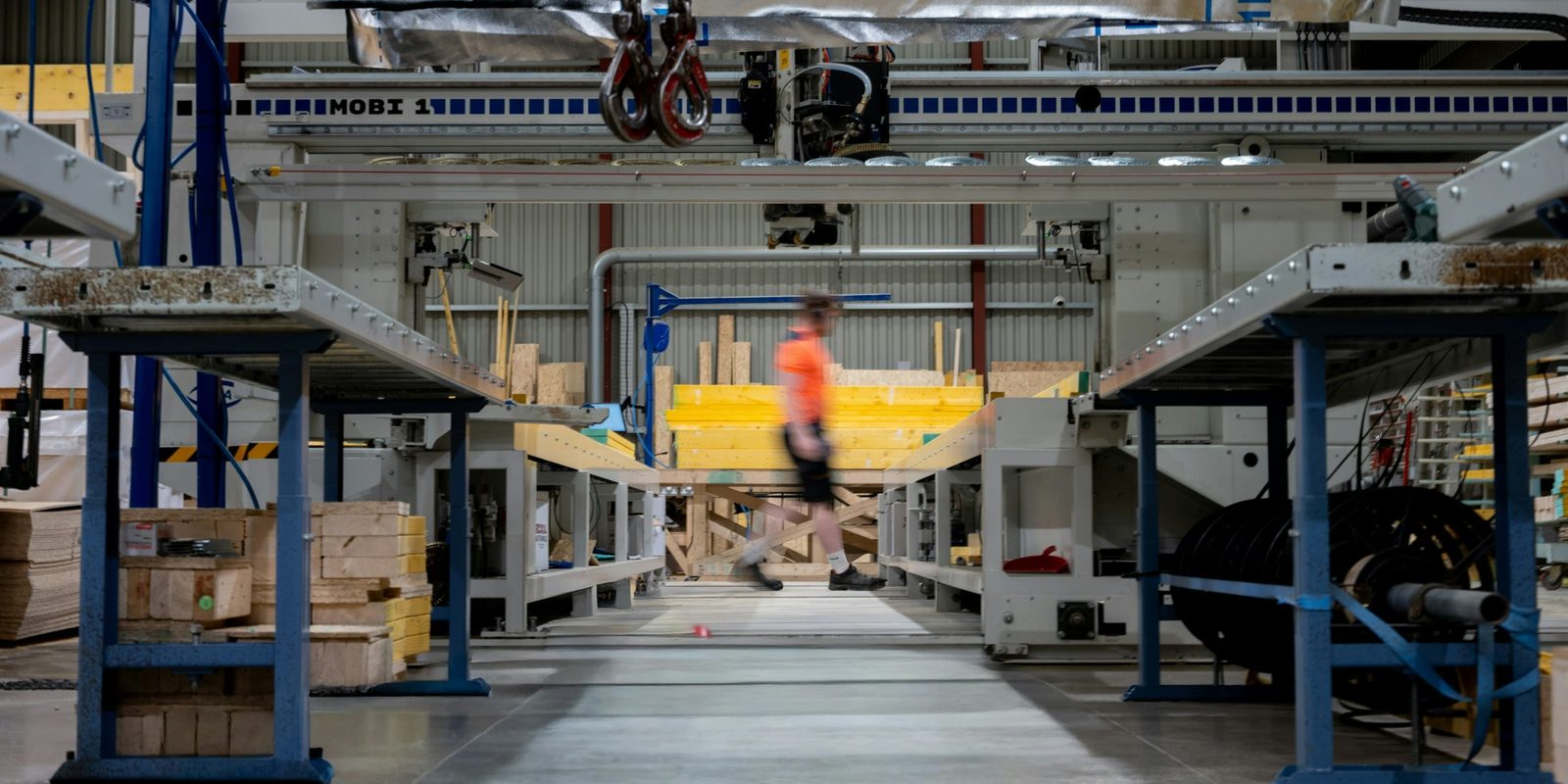 A worker in an orange shirt walks past roller tables under a large gantry machine in a modern wood products plant