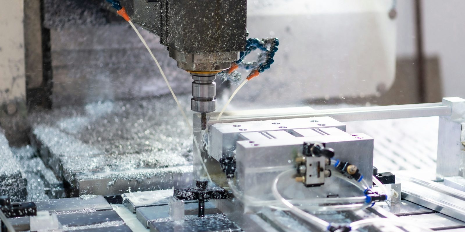 A CNC milling machine cuts an aluminum block, with coolant lines and metal chips around the spindle