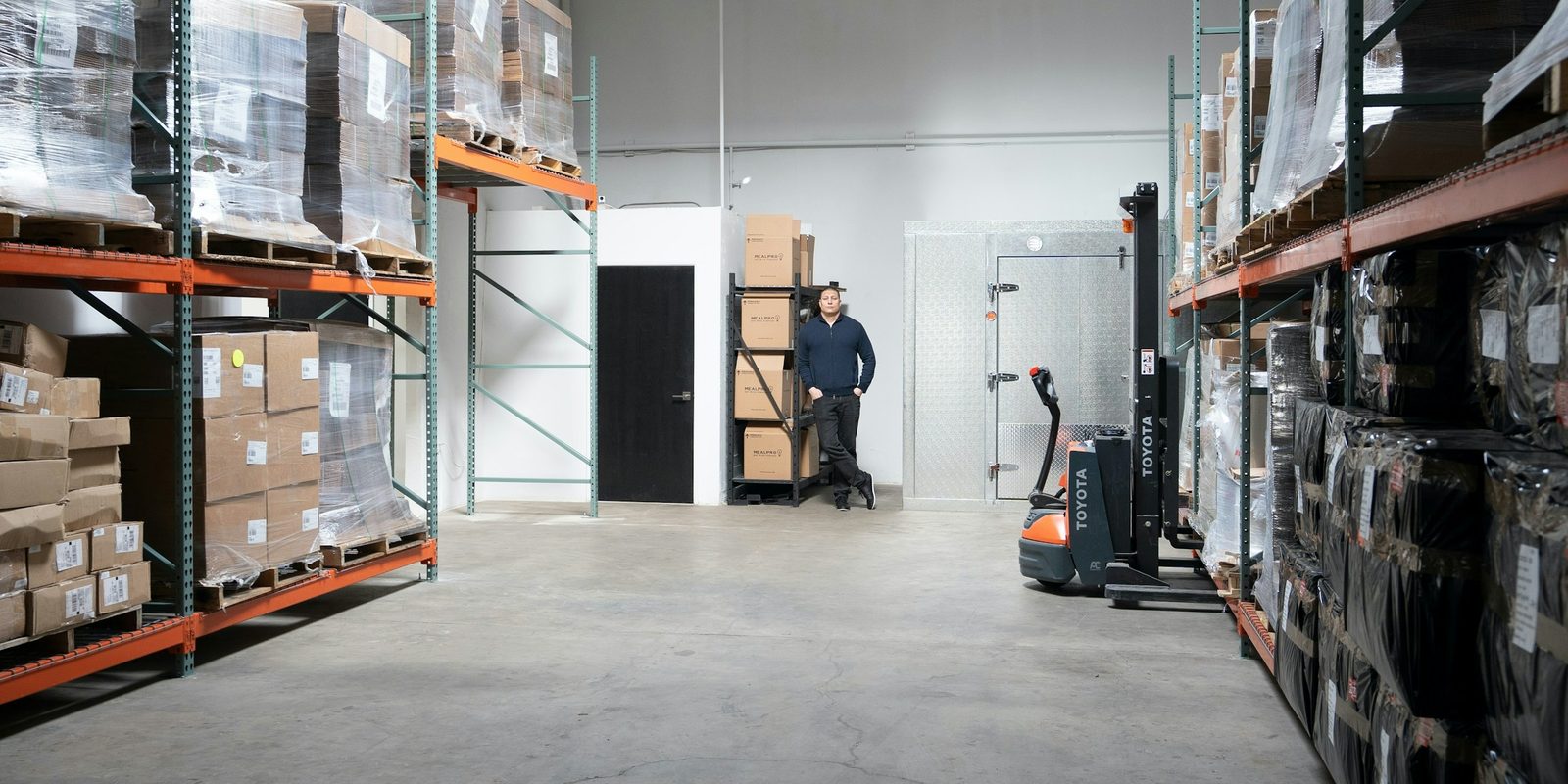 A bright warehouse with pallets of boxed stock on orange racks, a pallet truck and a man standing by the far wall