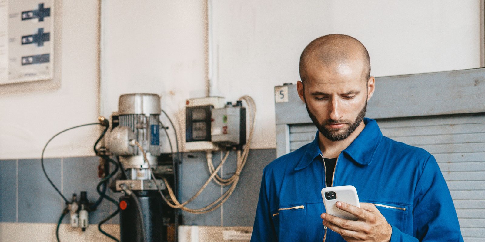 Technician in blue coveralls reads his smartphone beside wall-mounted machinery in a bright workshop