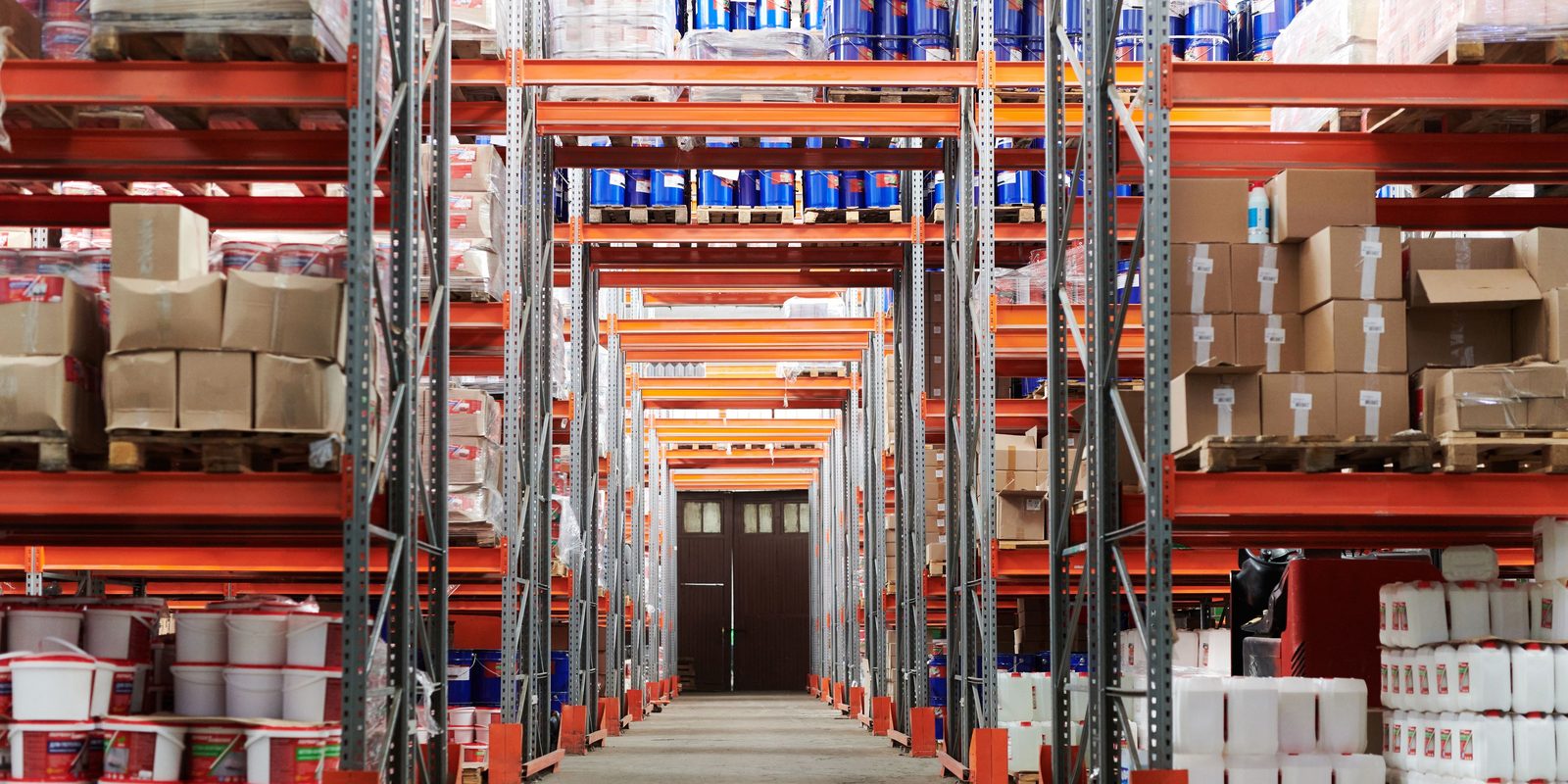 A long aisle of orange pallet racking stocked with buckets, canisters and cartons in a distribution warehouse