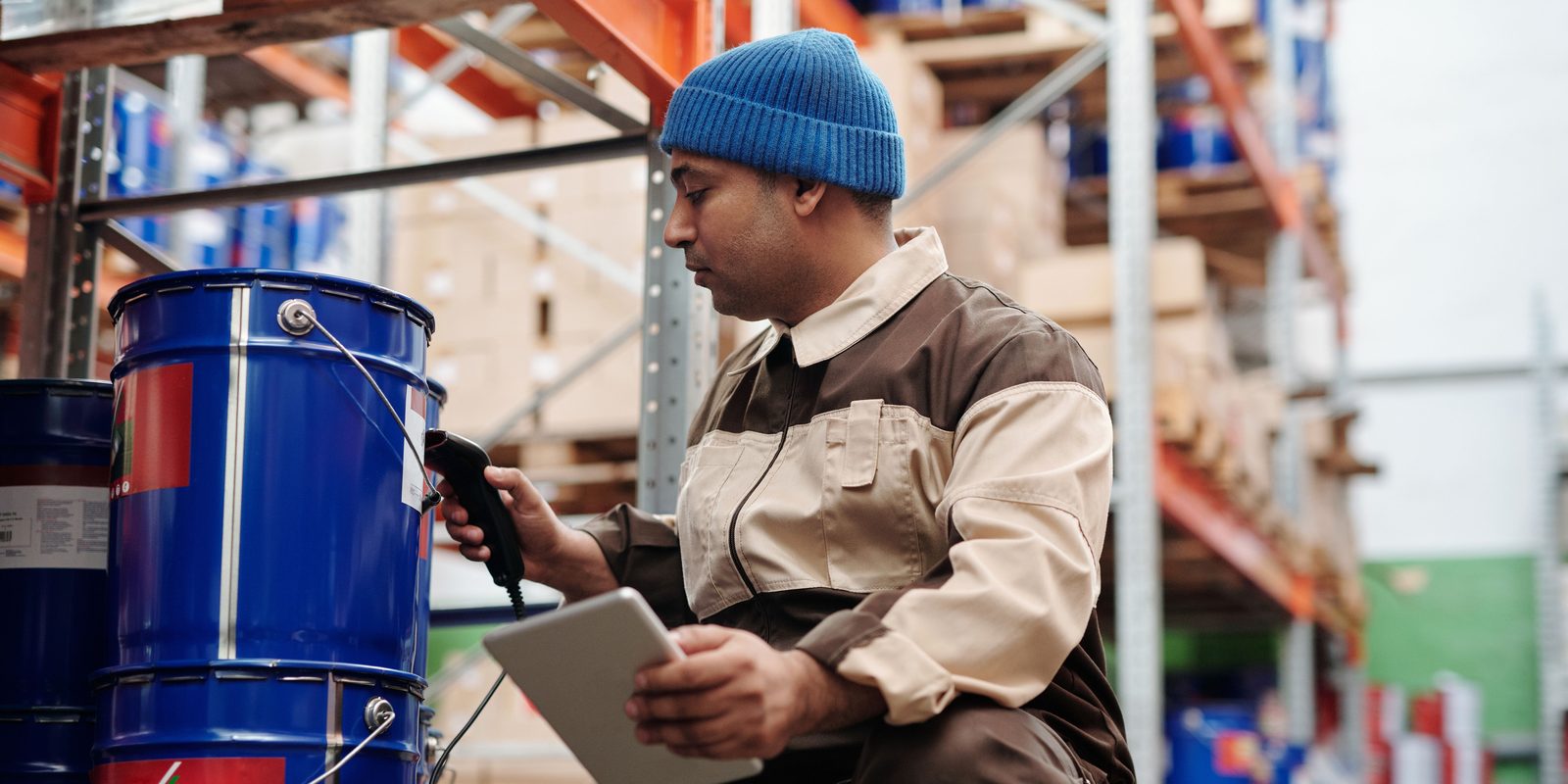 A warehouse worker scans the label on a paint drum and records it on a tablet beside pallet racking