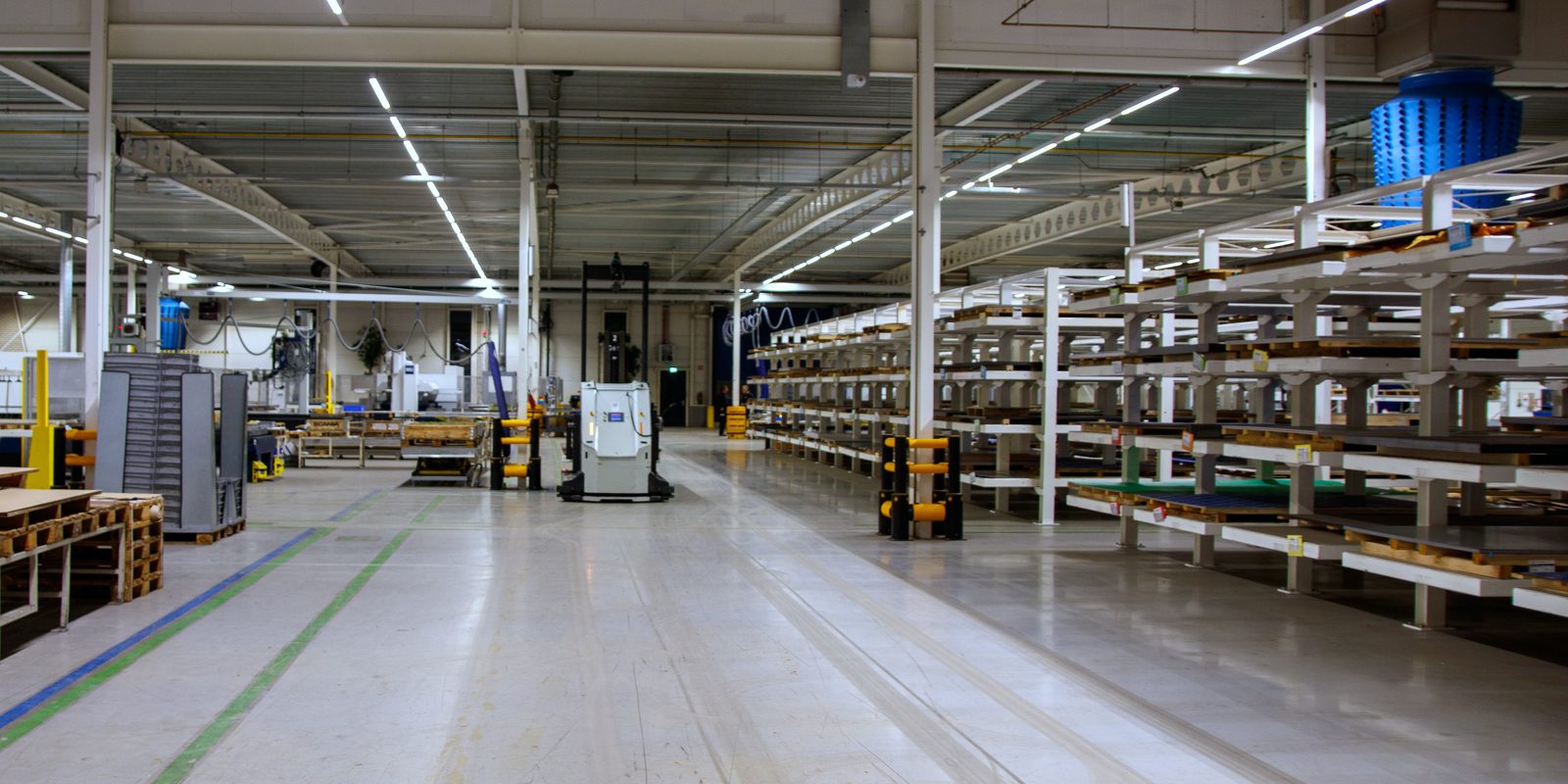 Wide, bright factory aisle with parts racking on one side, machines on the other and an automated cart in the lane