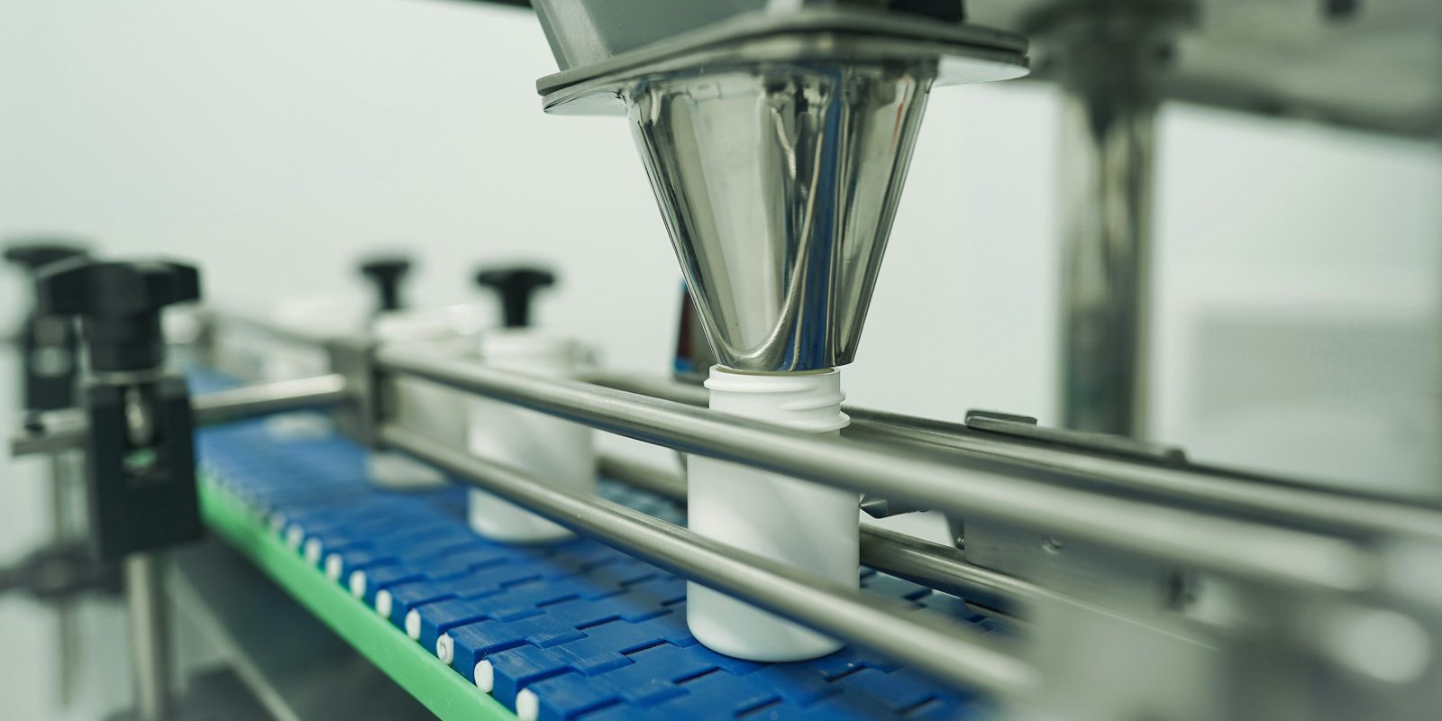 White bottles move along a blue conveyor under a stainless filling funnel on an automated line