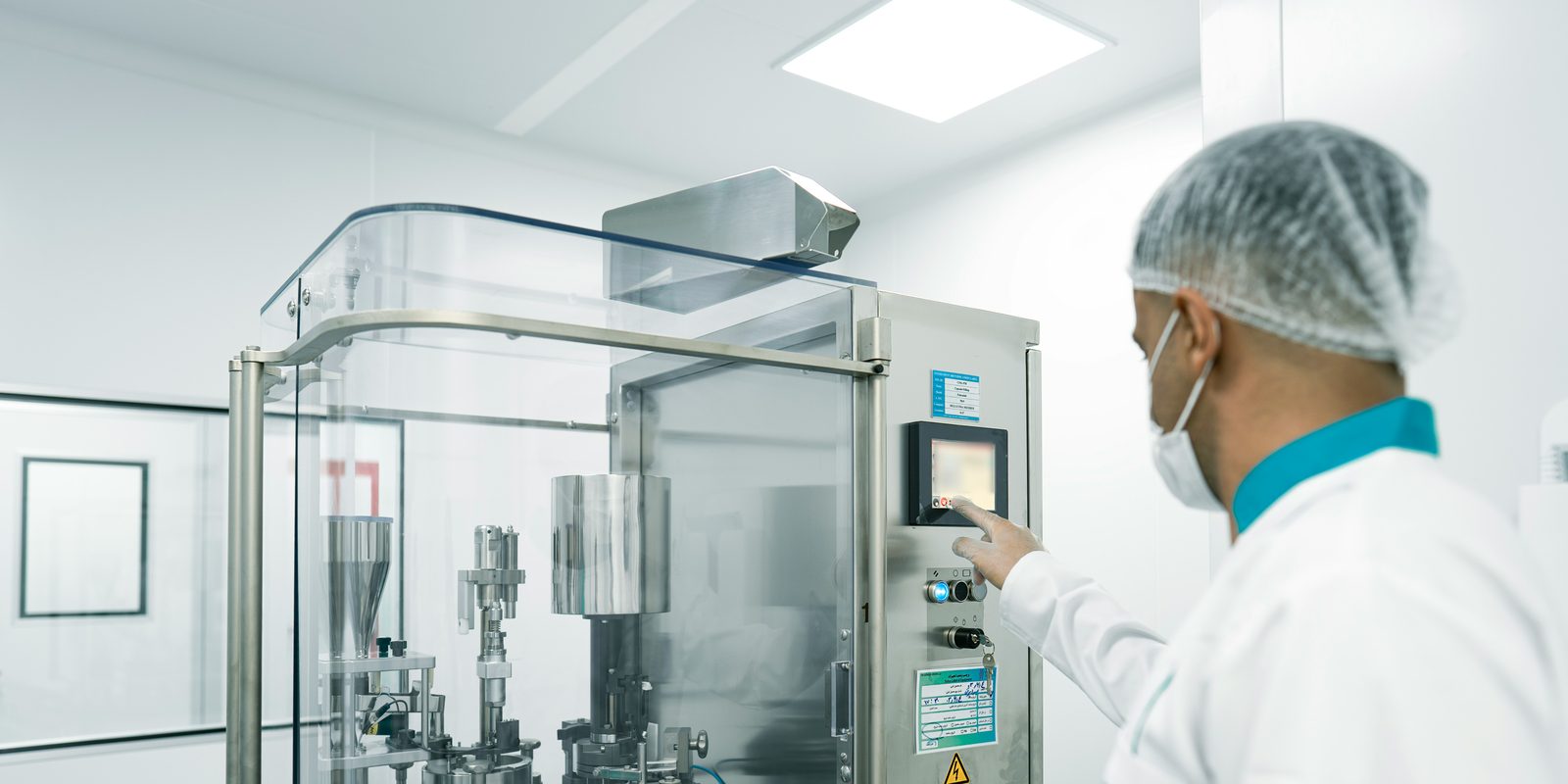 Operator in a hairnet and lab coat taps the touchscreen of a capsule filling machine in a bright cleanroom