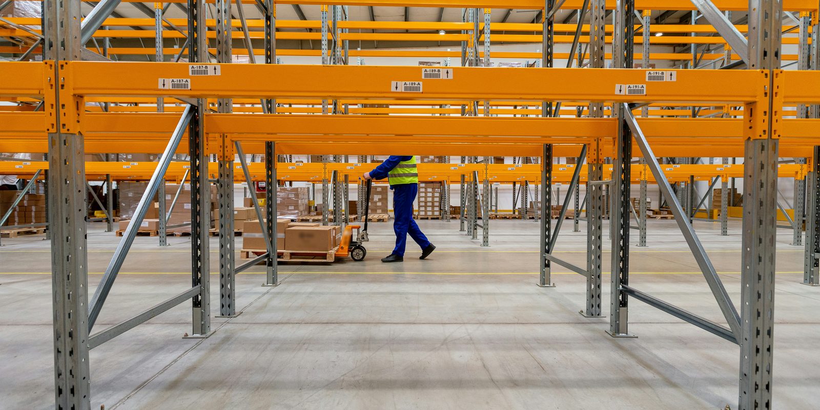 A worker pulls a pallet jack between rows of orange pallet racking with barcoded location labels in a bright warehouse