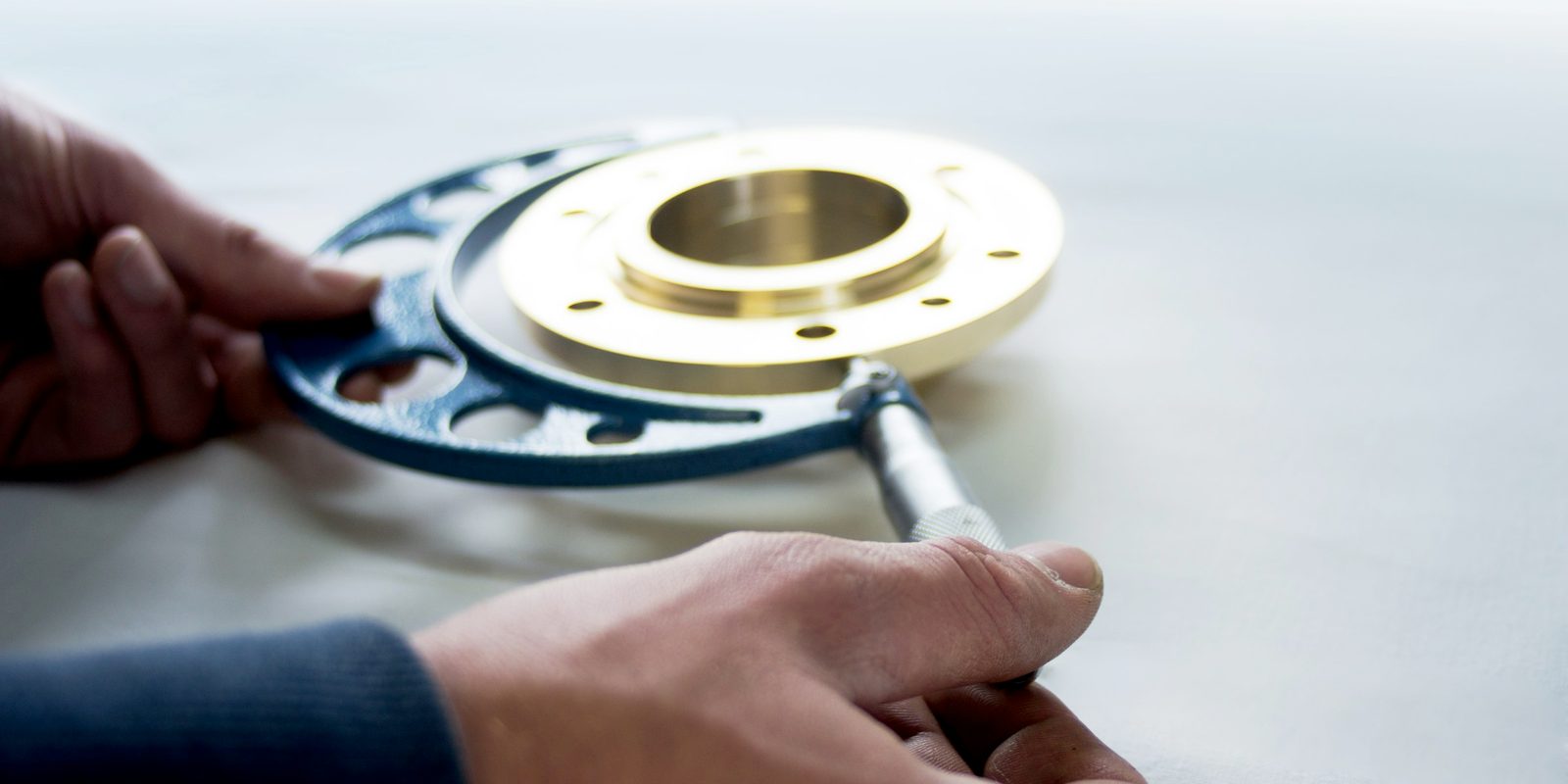 Hands measuring a brass flange with a large micrometer on a bright workbench