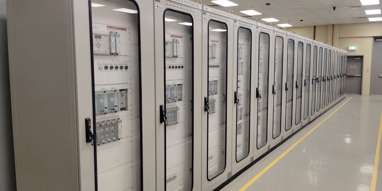 A long row of grey electrical cabinets with glass doors, each fitted with different breakers and relays, in a clean hall
