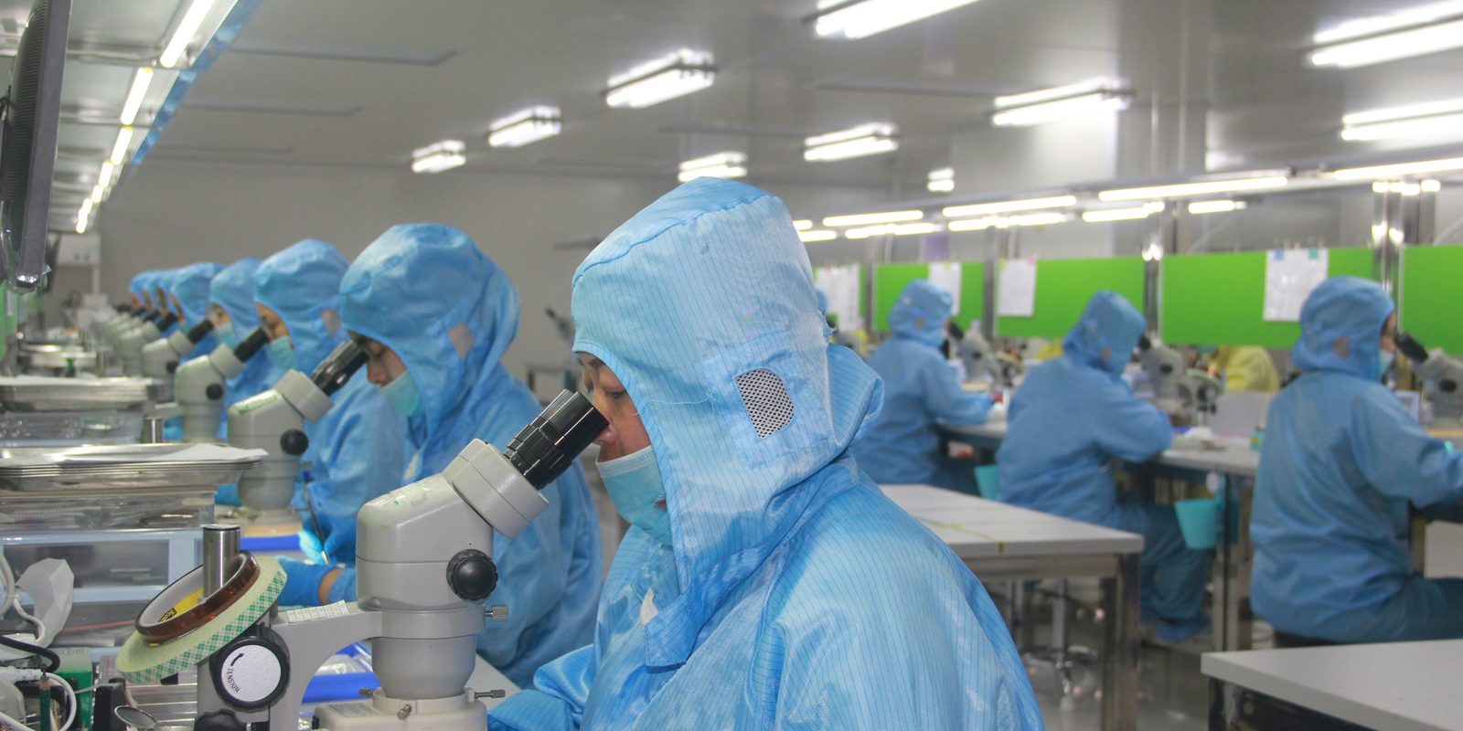 Technicians in blue cleanroom suits and hoods inspect small parts under stereo microscopes at long benches in a bright production room