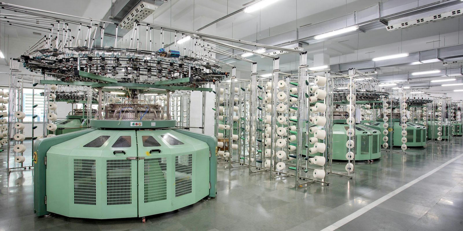 Rows of pale green circular knitting machines with white yarn cones on tall creels in a bright, clean factory hall